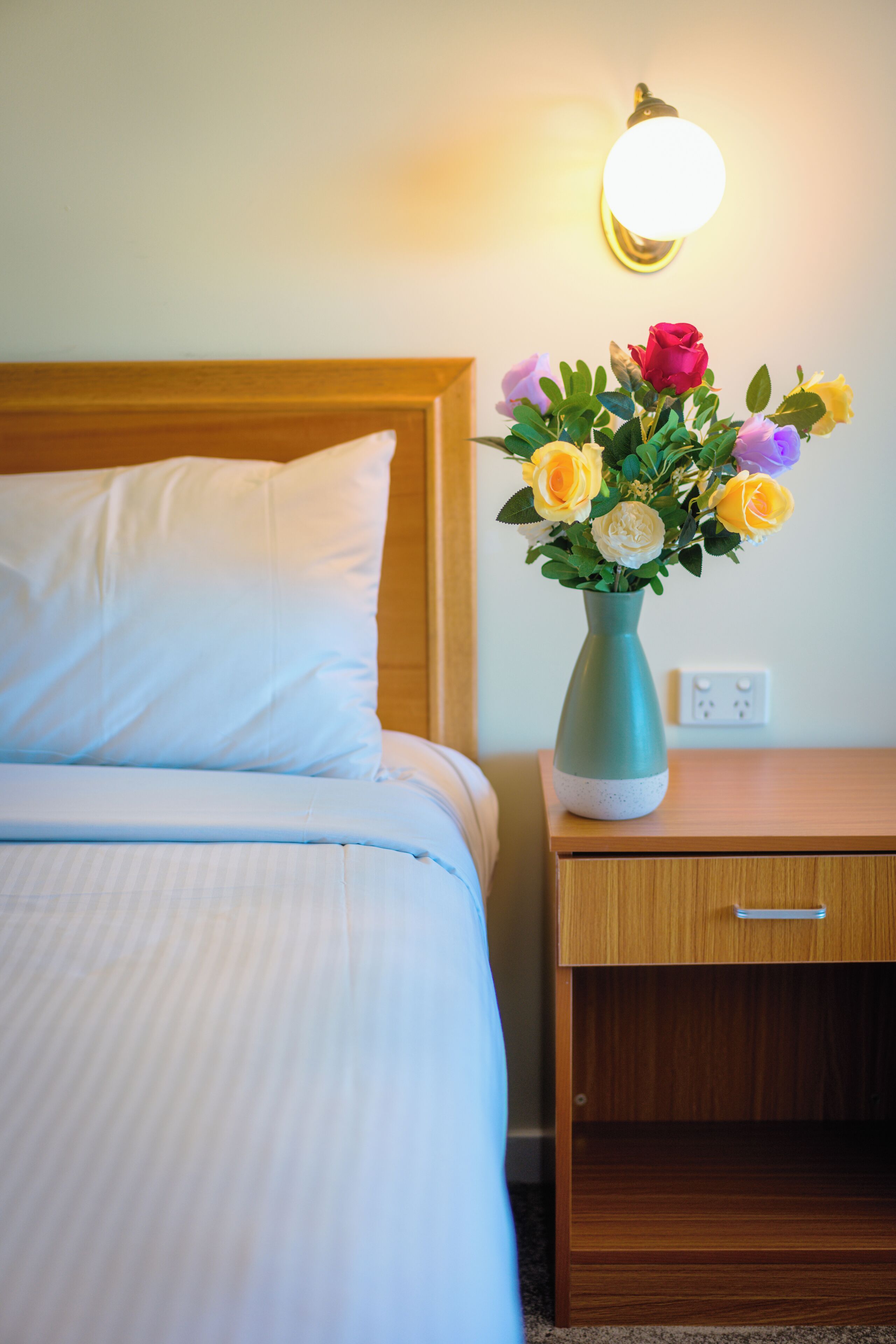 A neatly made bed with a white pillow and a teal vase with flowers on a wooden nightstand.