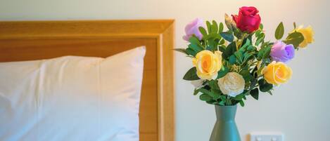 A neatly made bed with a white pillow and a teal vase with flowers on a wooden nightstand.