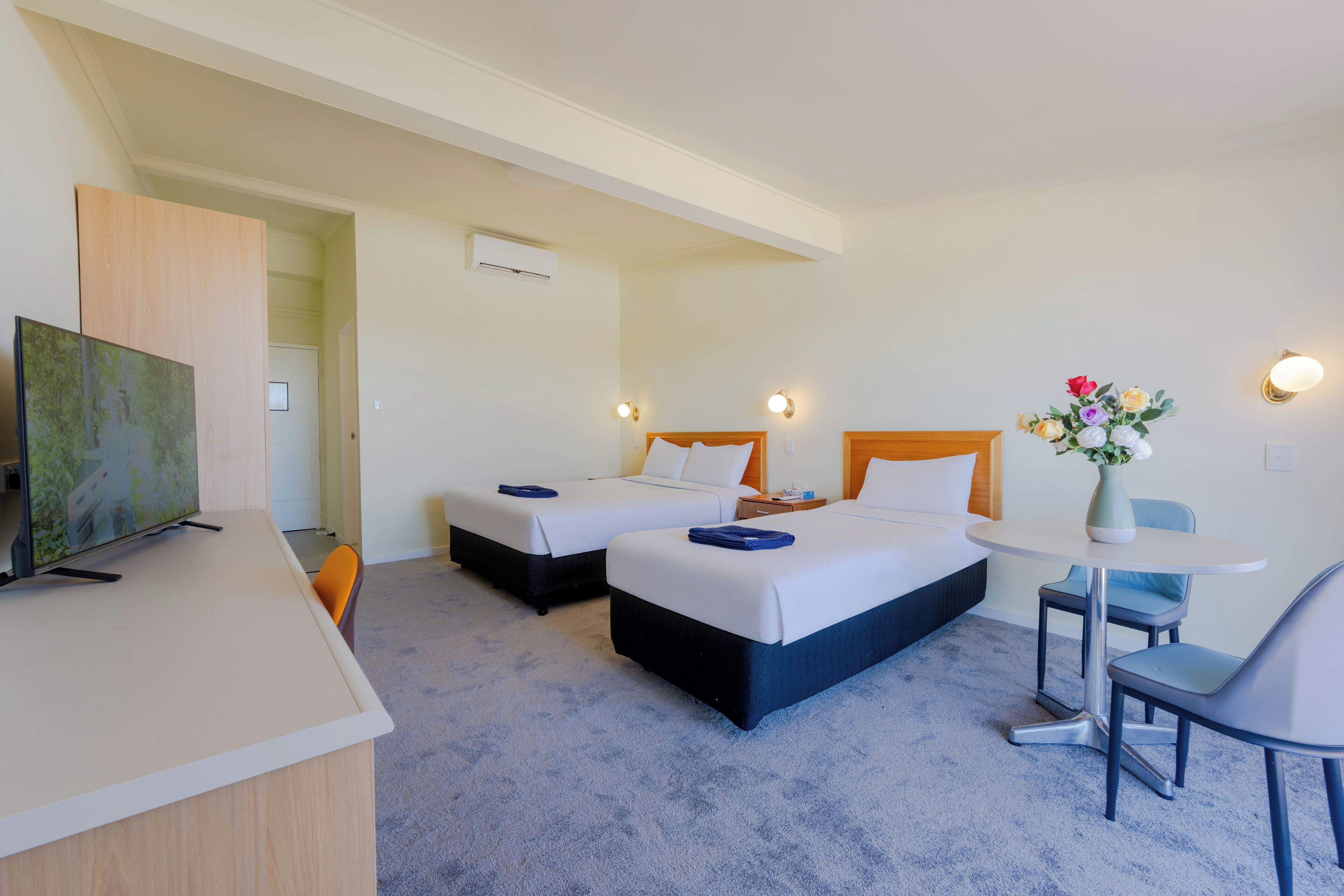 A hotel room with two beds, a desk, a chair, a small table, and a vase with flowers.