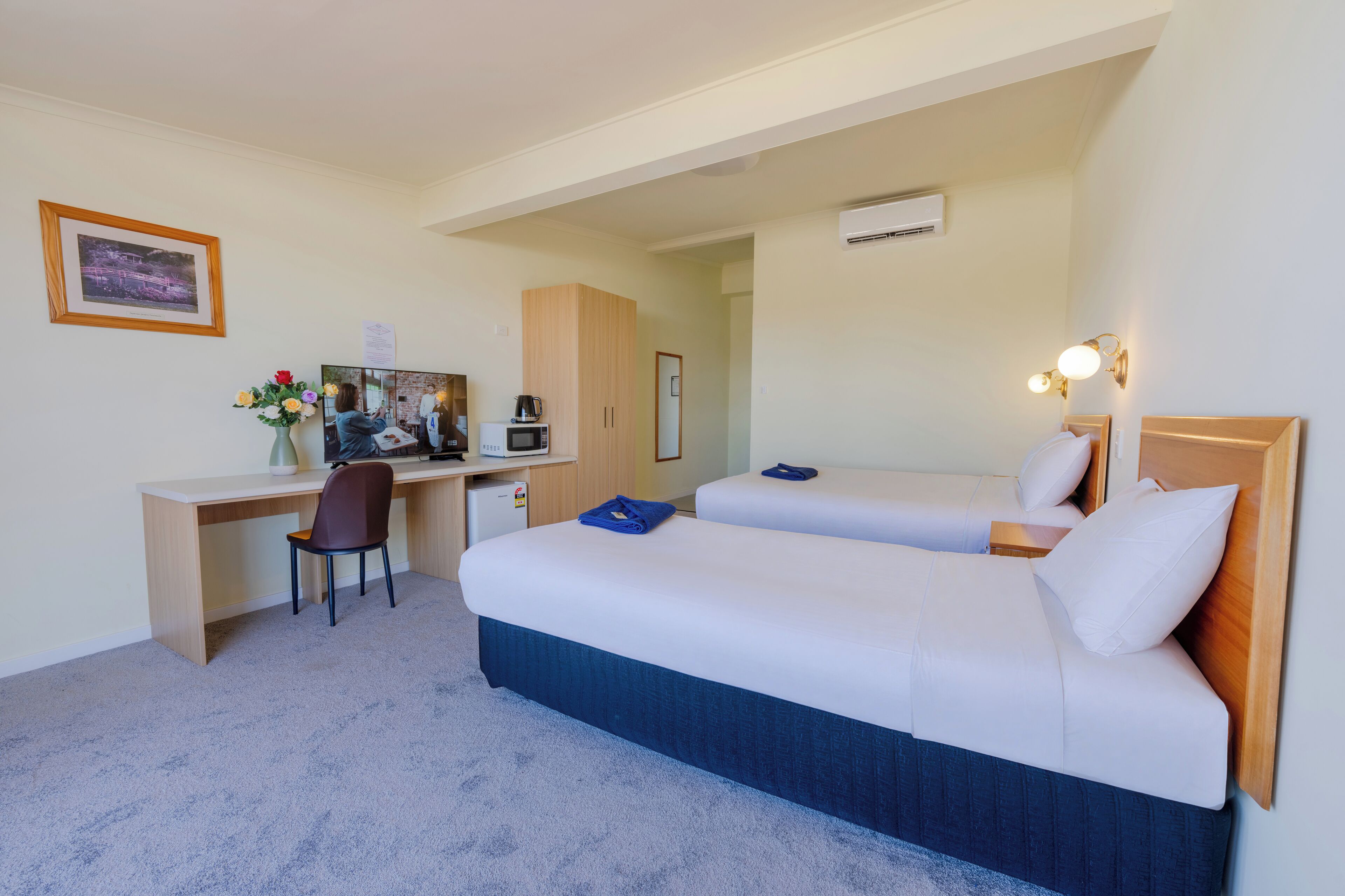 Une chambre d’hôtel équipée d’un bureau, de deux lits, d’une chaise, d’un petit coin cuisine et d’un tableau encadré au mur.