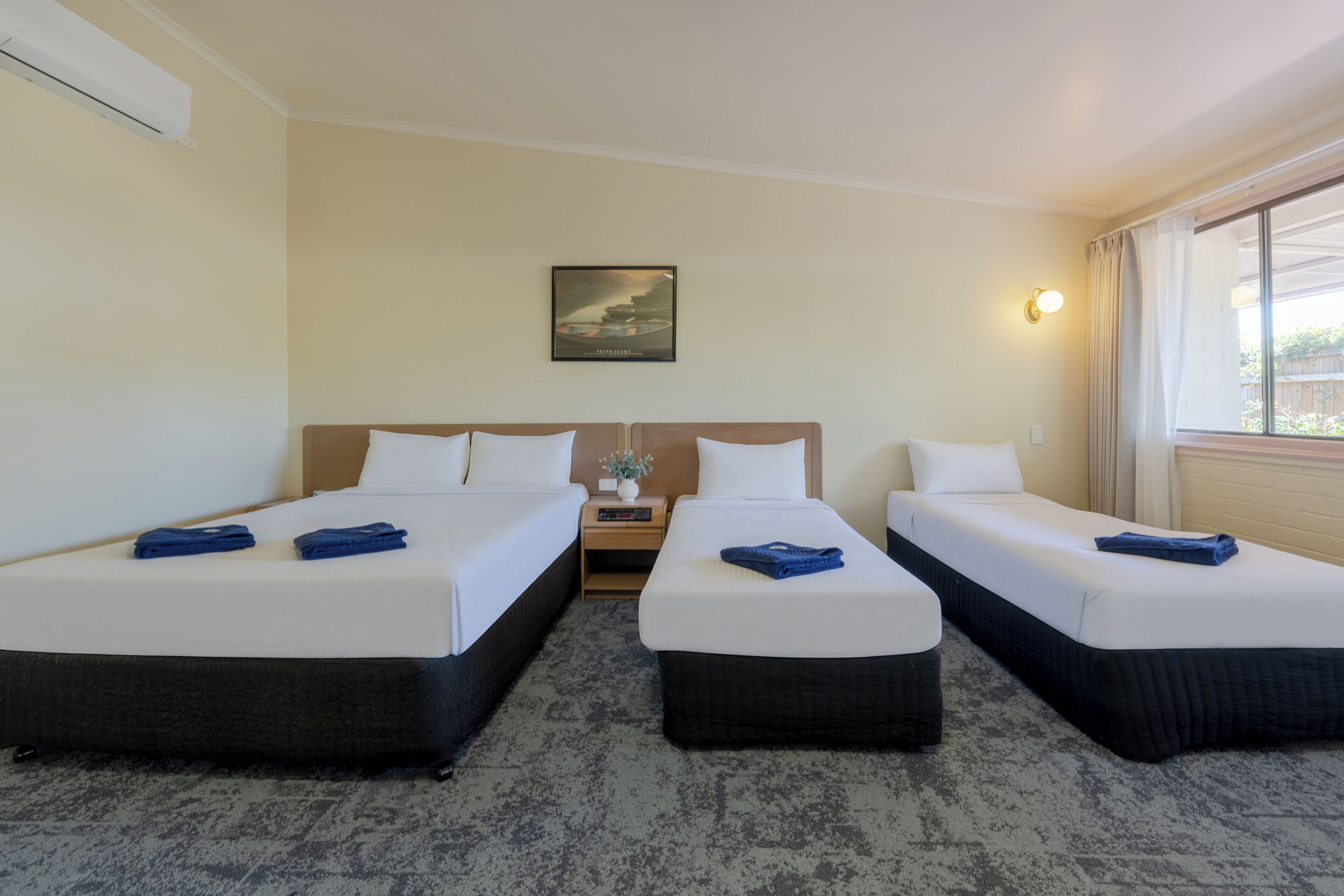 Une chambre d’hôtel avec deux lits, une table de chevet, un tableau accroché au mur et une fenêtre avec des rideaux.
