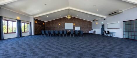 A large, open room with a high ceiling, a brick wall, and a carpeted floor. There are rows of chairs and a projector screen.
