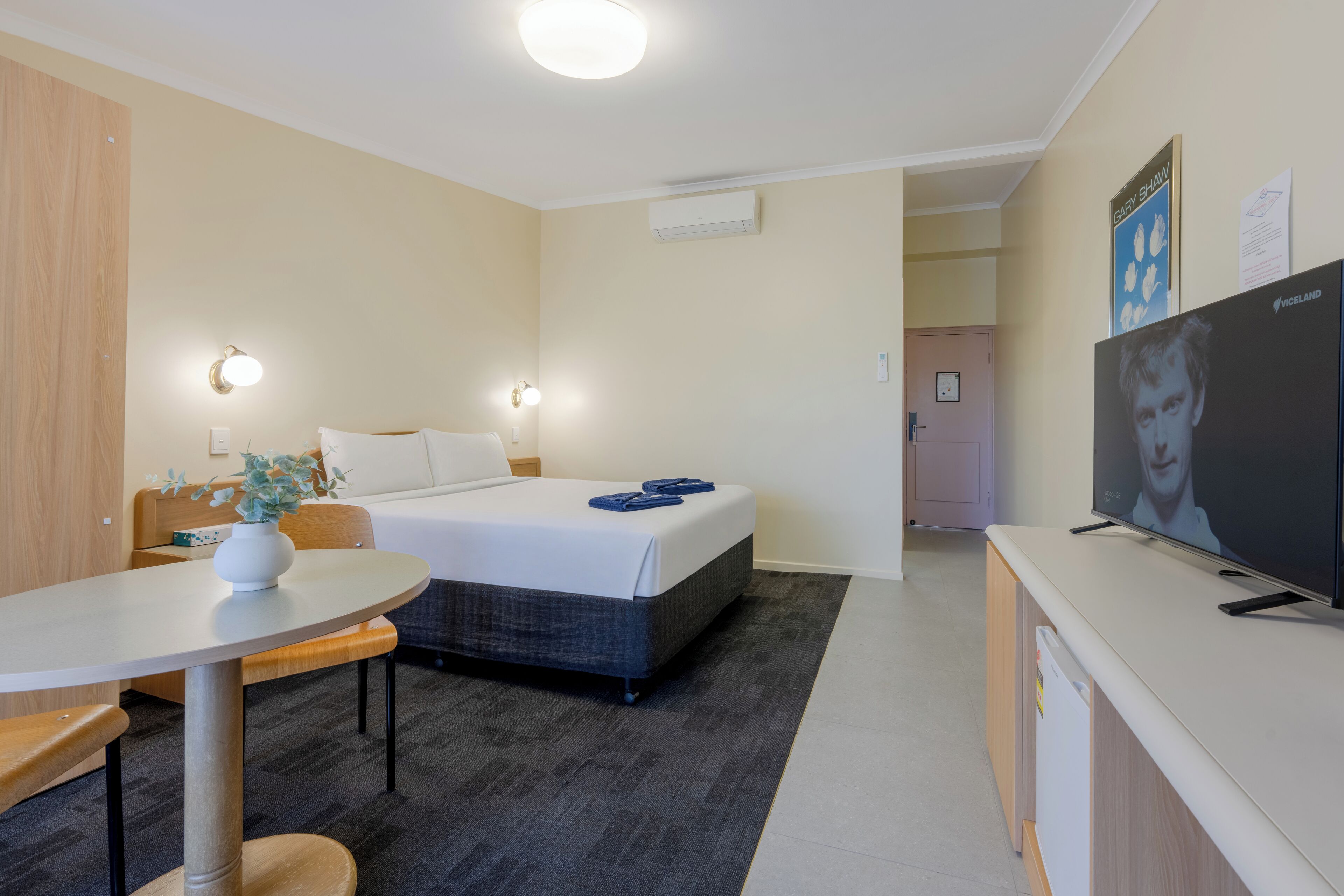 Habitación de hotel con cama, escritorio, silla, televisor y una mesita con un florero.