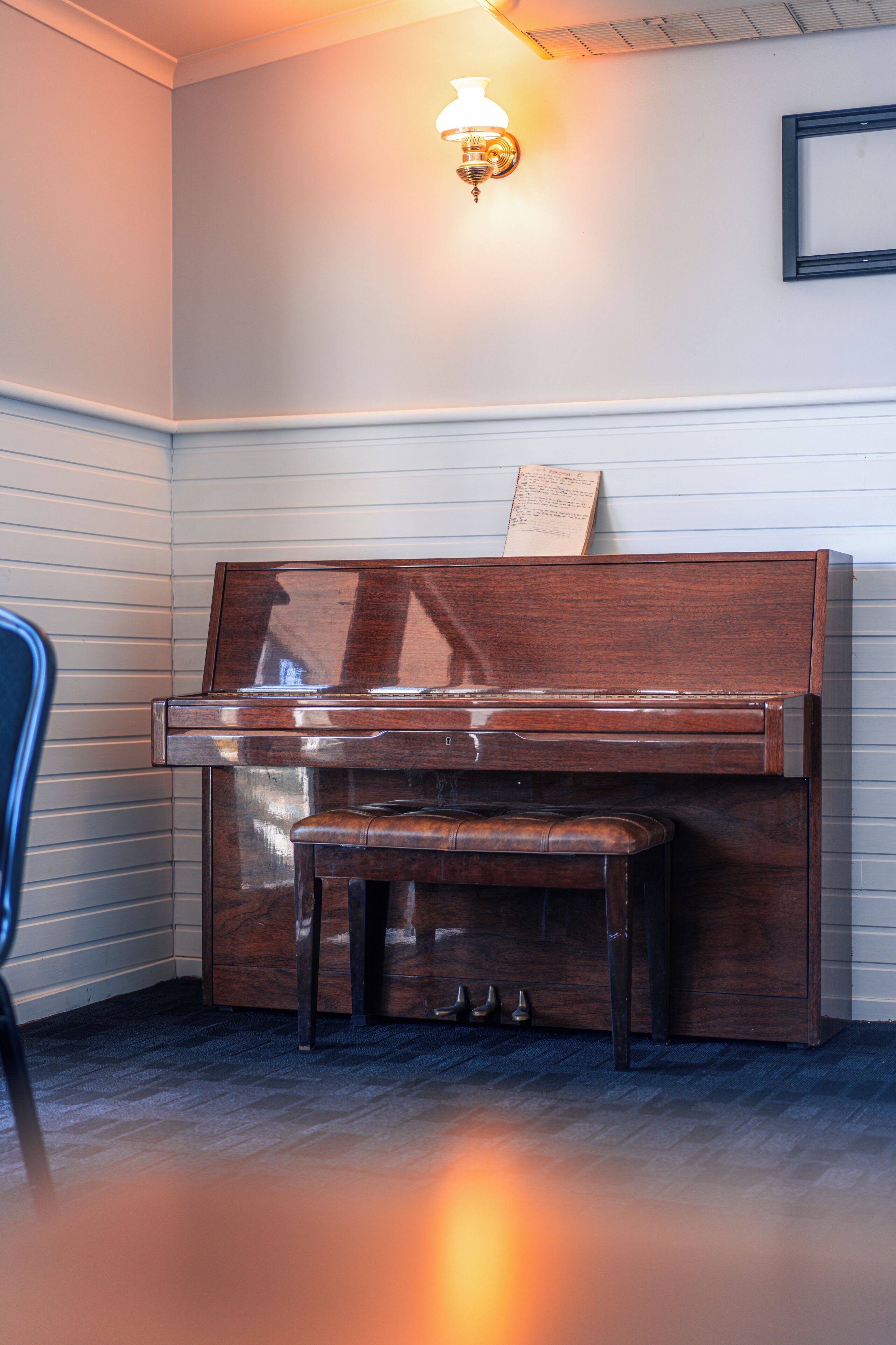 A piano with sheet music on top, a bench, and a chair in a corner room with a light fixture on the wall.