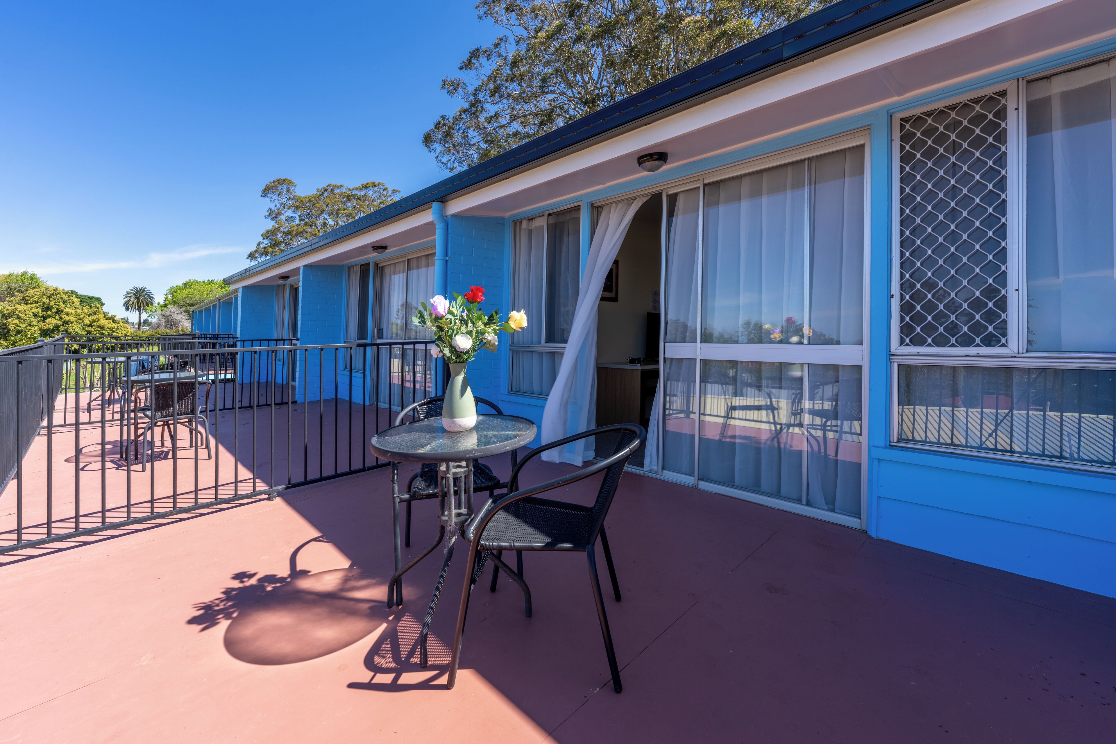 Un motel bleu avec un balcon, une table avec des chaises et un vase de fleurs.