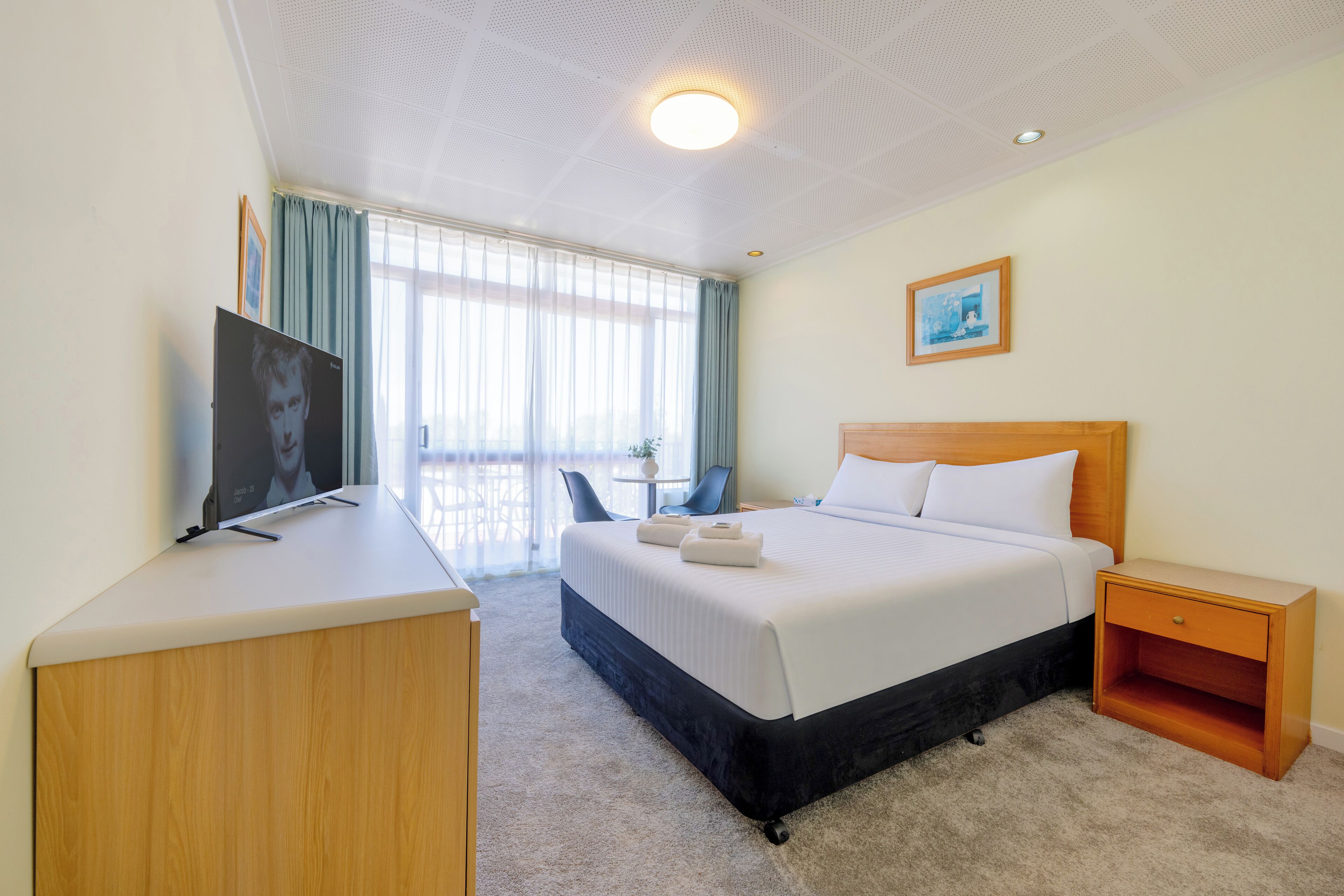 A hotel room with a bed, a desk, a chair, a nightstand, a TV, and a window with curtains.