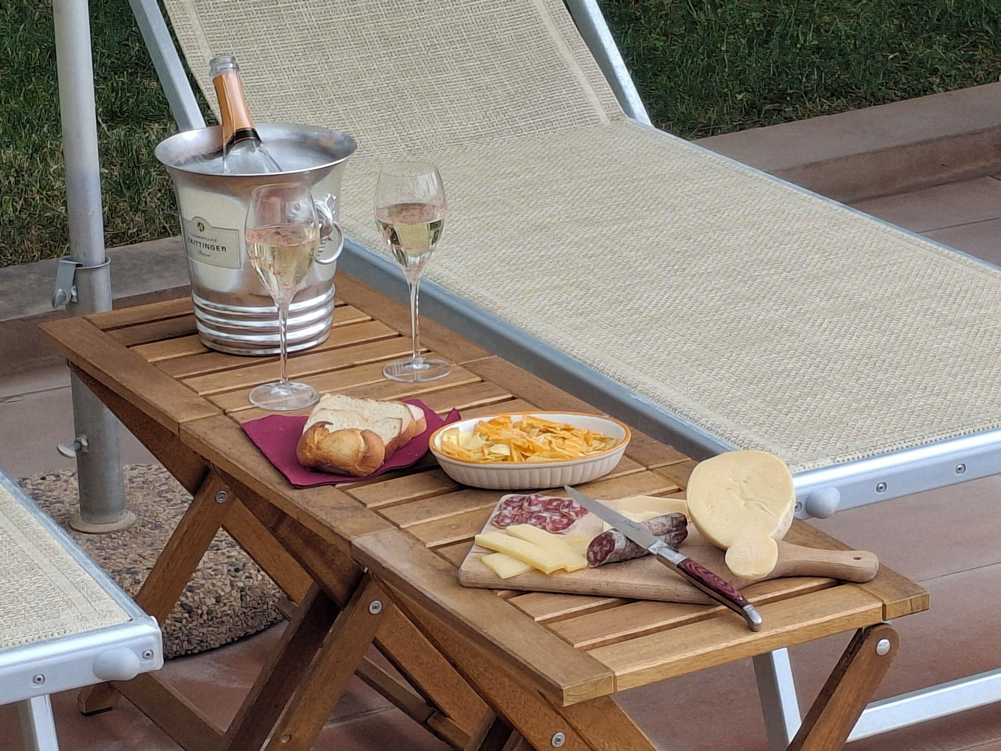 A wooden table with a bottle of champagne, two glasses, a bowl of chips, a cheese board, and a knife.