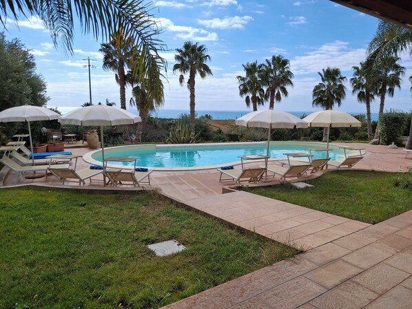 A pool area with lounge chairs and umbrellas, surrounded by palm trees and a view of the ocean.