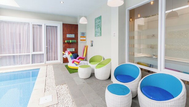 A poolside area with a clear view of the pool, wicker chairs with blue cushions, a colorful inflatable unicorn ride, and a large sliding glass door. - Omah Manis Taman Siswa (Yogyakarta)