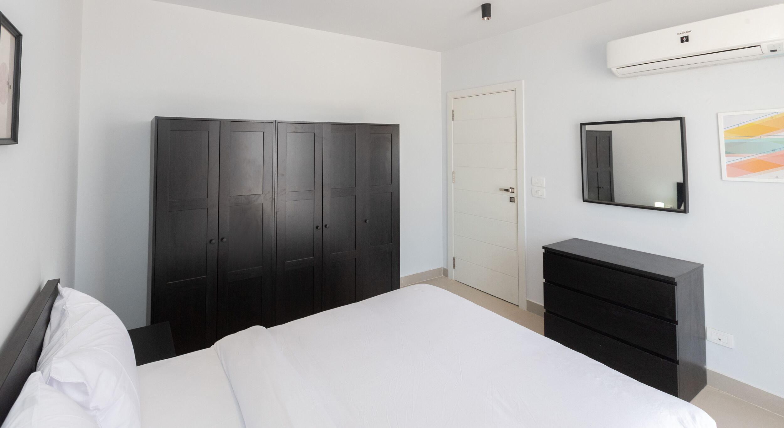 A bedroom with a large bed, a wardrobe, a dresser, a mirror, and an air conditioning unit.