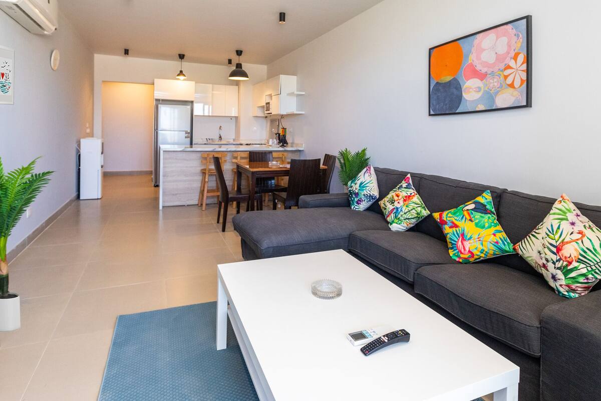 A modern living room with a grey sofa, a white coffee table, and a dining area in the background.