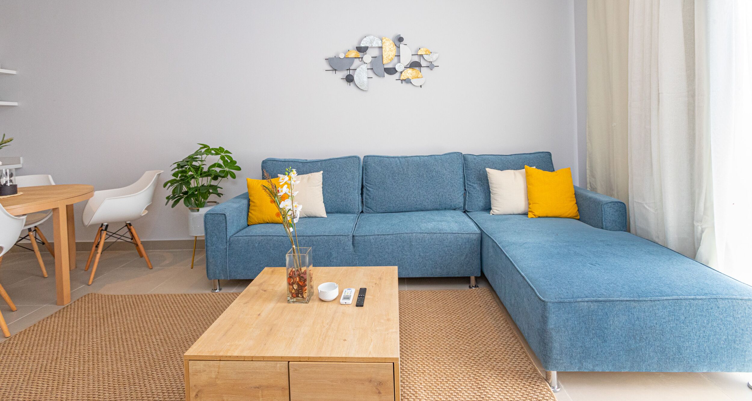 A modern living room with a blue sectional sofa, a wooden coffee table, and a dining area with white chairs.