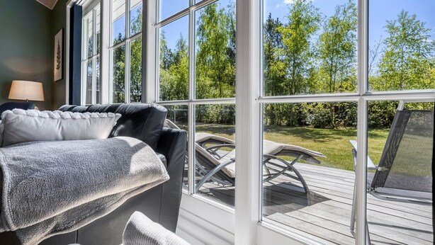 A sunlit room with a large window, a sofa with a blanket, and a view of a deck and greenery outside.