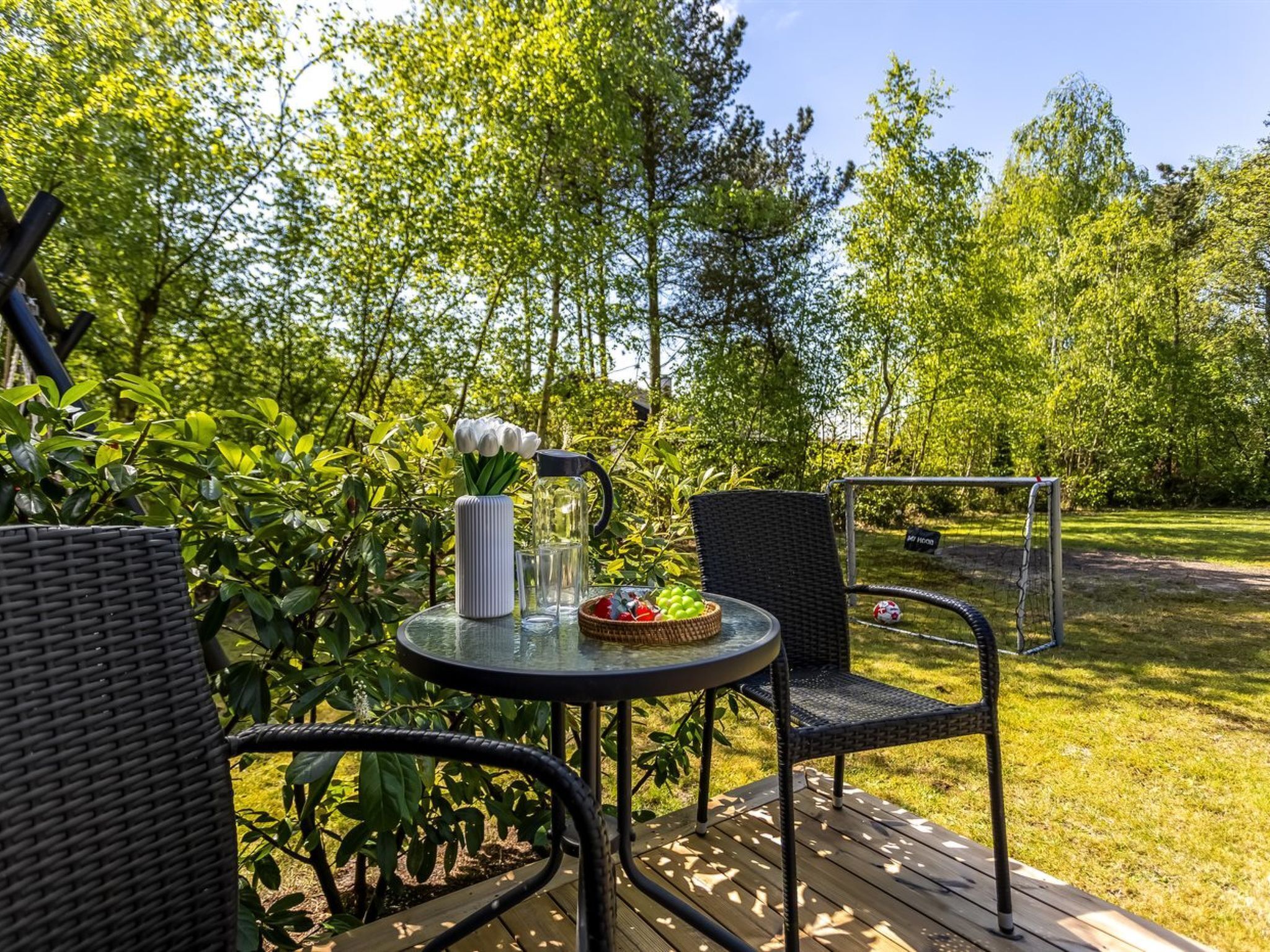 Un patio al aire libre con una mesa redonda, dos sillas de mimbre, una silla de metal y una jarra de vidrio con tulipanes.