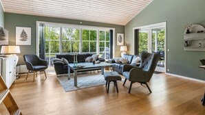 A spacious living room with a high ceiling, large windows, and a view of greenery outside.