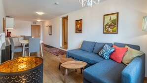 Ein modernes Wohnzimmer mit einem blauen Sofa, einem Holztisch und einer Chandelieber. Es gibt eine Essecke mit Tisch und Stühlen, und im Hintergrund eine Küche.