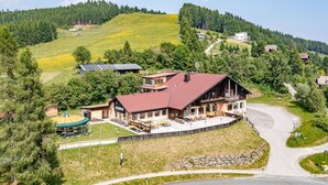 A large lodge with a red roof, a covered outdoor seating area, and a view of a winding road and forested hills. - Nice apartment for 5 people with WIFI, balcony and panoramic view (Frauenstein)
