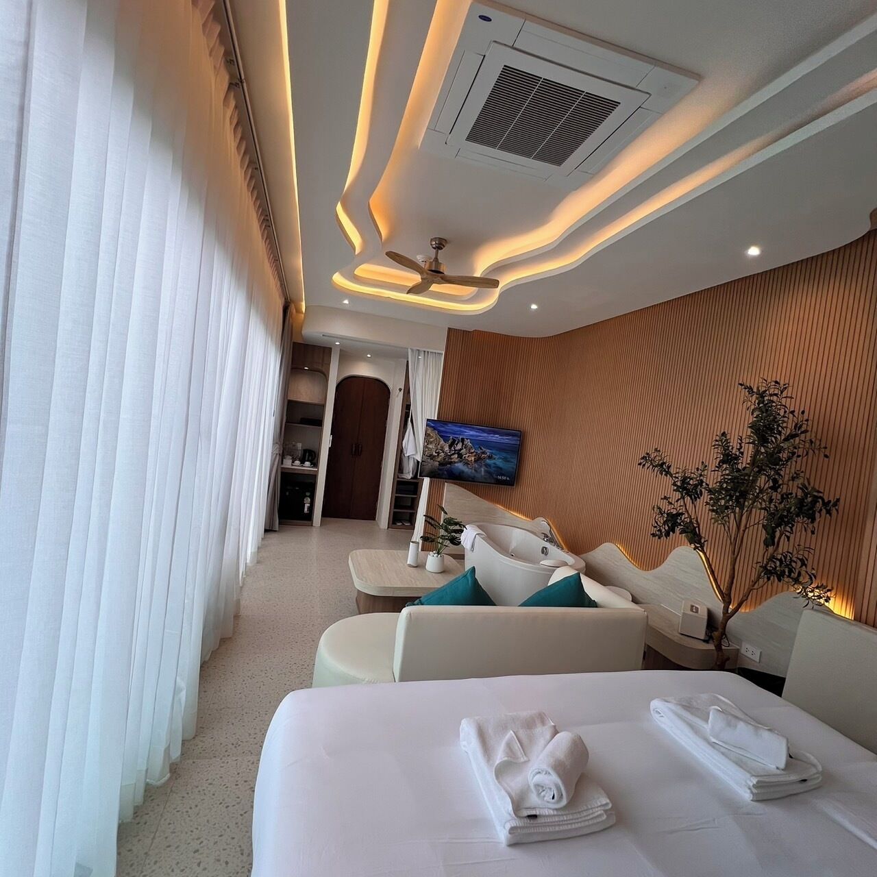 A modern hotel room with a large bed, a seating area with a sofa and armchairs, a flat-screen TV, and a ceiling fan.