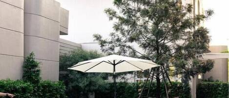 Zona de piscina con dos sillones blancos y una sombrilla grande, rodeada de follaje y un edificio moderno.