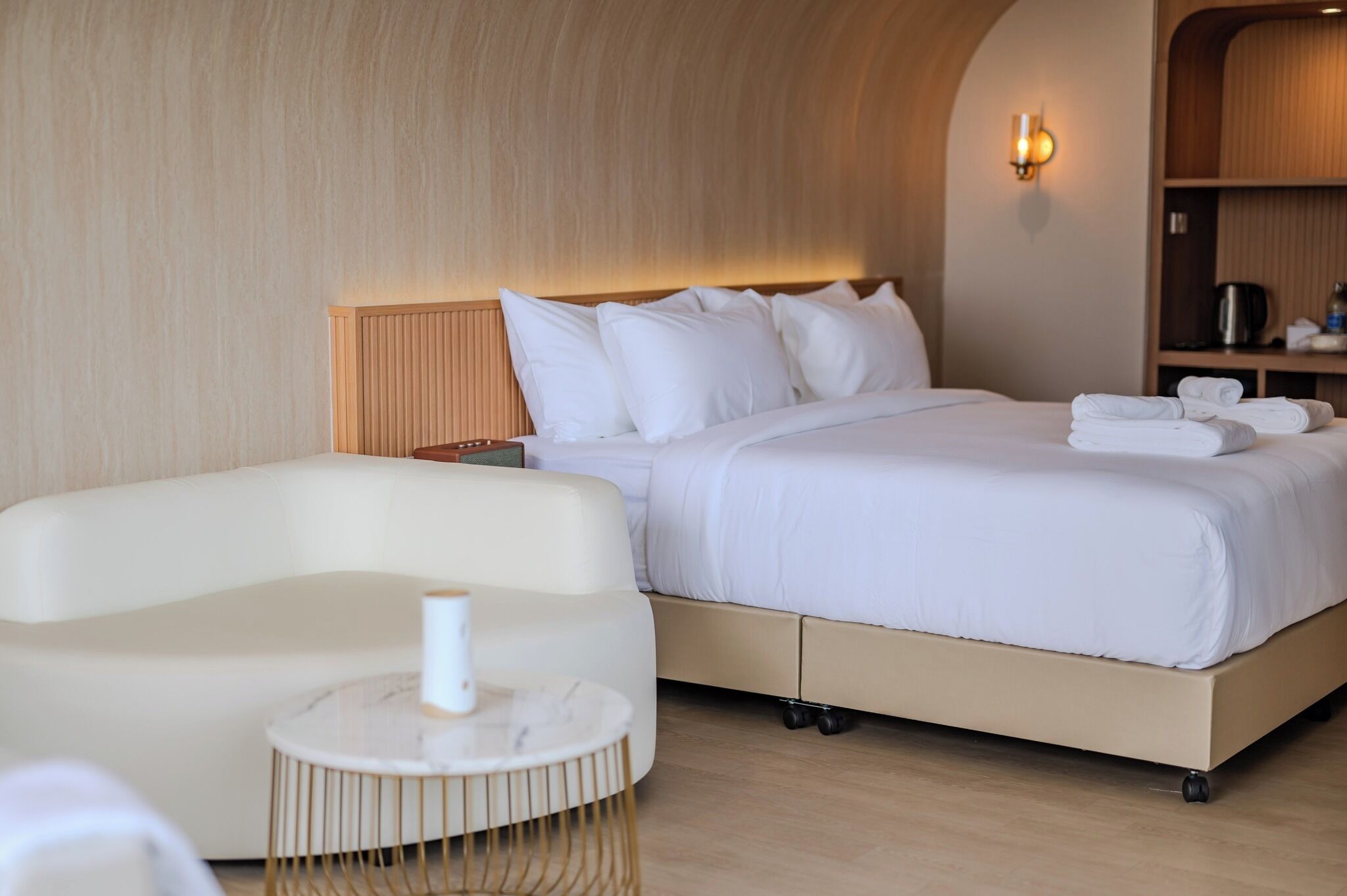A modern hotel room with a bed, a sofa, a small round table, and a wall-mounted shelf with a kettle.