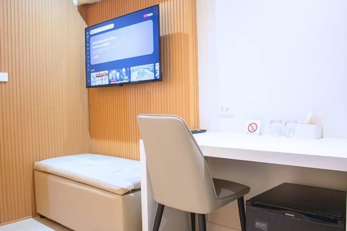 A hotel room with a television mounted on the wall, a desk with a chair, a small bedside table, and a trash bin.