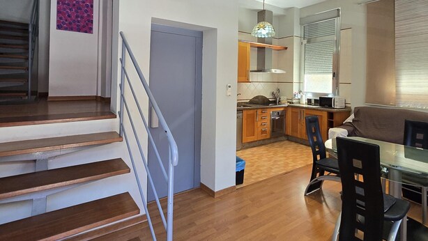 Una cocina moderna con armarios de madera, un comedor con mesa de vidrio y una escalera con barandillas de metal.
