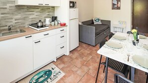 A compact kitchen with a sink, stove, and a small refrigerator. There is a dining table with chairs and a sofa in the adjacent living area. - Nice apartment for 5 people with A/C, pool, WIFI and TV (Castelveccana)