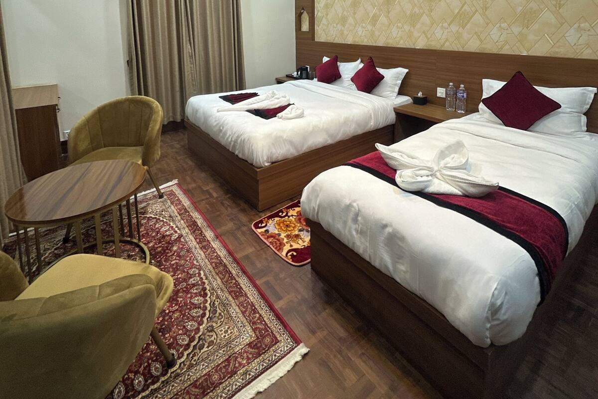 A hotel room with two beds, a wooden headboard, a small table, a chair, and a red and gold patterned rug.