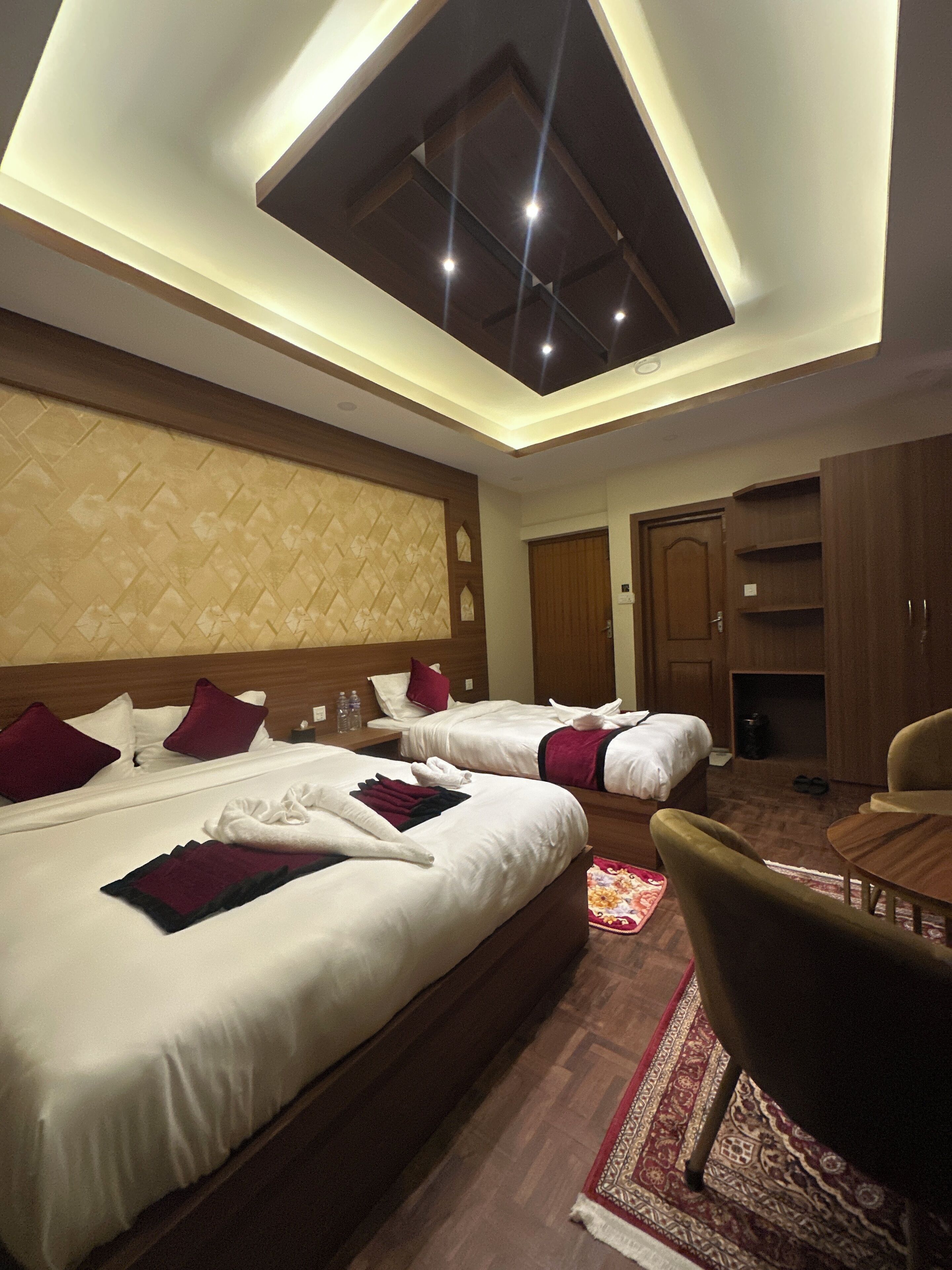 A hotel room with two beds, a wooden headboard, a ceiling with recessed lighting, a wooden wardrobe, and a small table with a chair.