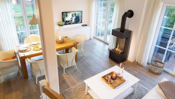 A modern living room with a fireplace, a dining area with wicker chairs, and a television on the wall. - Ferienhaus Strandhafer (Wangerooge)