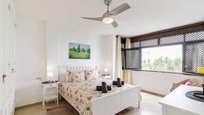 Een slaapkamer met een bed, nachtkastjes, een plafondventilator, een raam met uitzicht op groen, en een schilderij aan de muur.