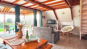 Ein geräumiger Wohnbereich mit Holzbalkendecke, einem grauen Sofa, einem Korbstuhl und einem Esstisch mit Untersetzern und einer Flasche.