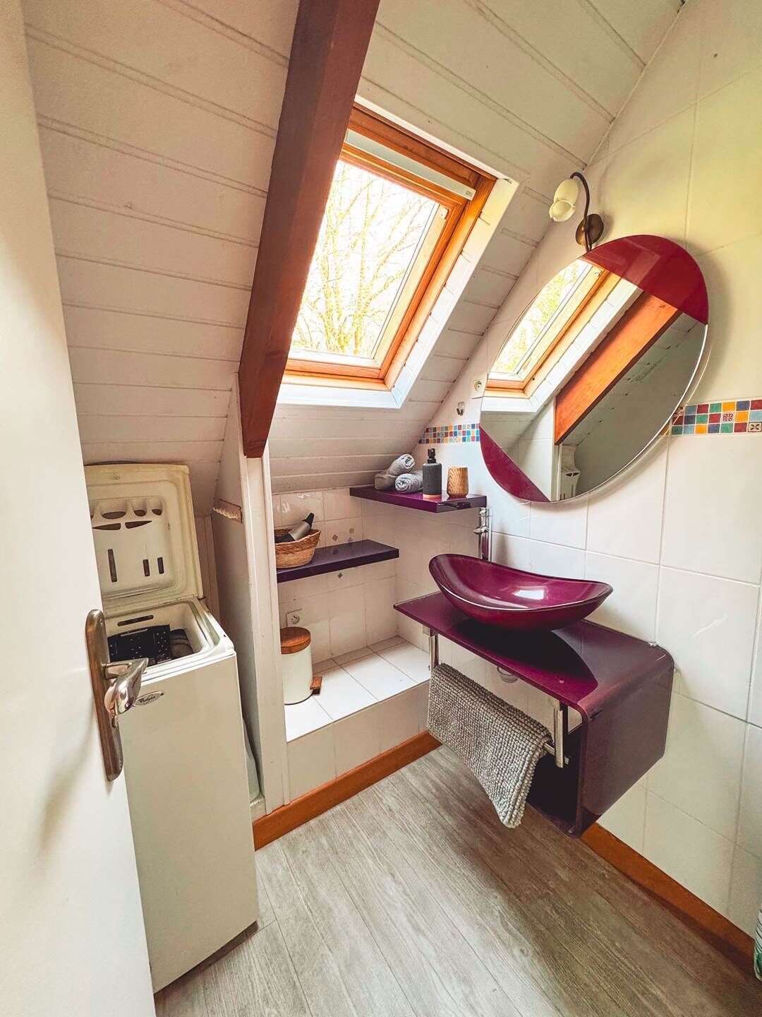 Een compacte badkamer met dakraam, een witte koelkast, een paars wastafel en een spiegel.