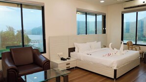 A modern bedroom with a large bed, a glass coffee table, a brown leather chair, and large windows offering a view of a body of water. - Praveg Rezort Silvassa (Silvassa)