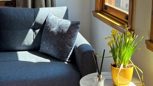 Ein Zimmer mit einem dunkelblauen Sofa, einem kleinen runden Tisch mit einem gelben Blumentopf und einem Fenster mit Blick auf Gebäude.