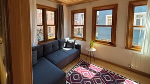Ein Zimmer mit Holzboden, einem blauen Sofa, einem gemusterten Teppich und großen Fenstern mit Blick auf Gebäude.