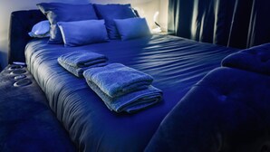 A neatly made bed with a blue blanket and pillows, a bedside table with a lamp, and a window with curtains. - The Floor - Ombre D'Ebene Loft & private Jacuzzi (Coignières)
