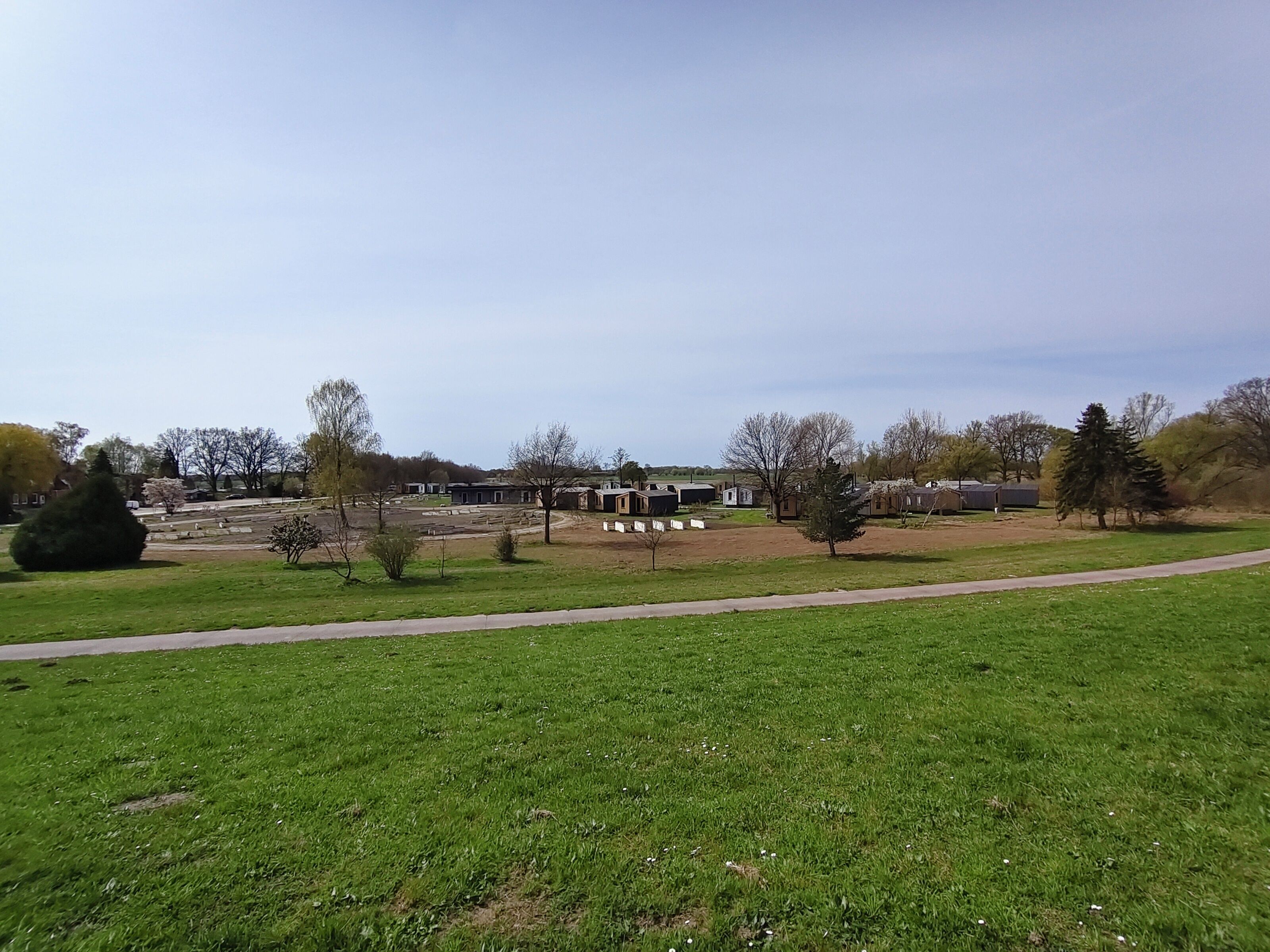 A grassy field with a paved path, a row of buildings, and trees in the background.