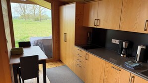 A compact kitchen with wooden cabinets, a black countertop, and a view of a grassy field through a large window. - MoJo's Tiny House on the Elbe (Bleckede)