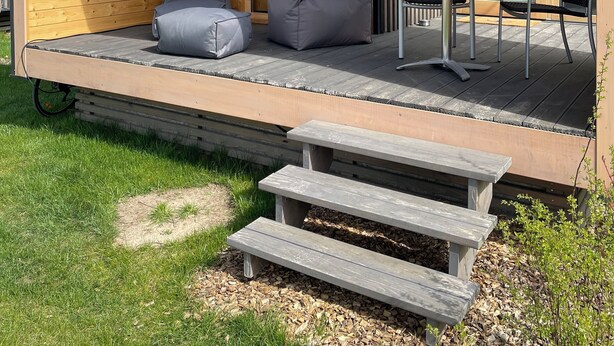 A modern wooden cabin with a covered porch, a large window with a view of a lake, and a dining area with outdoor furniture. - MoJo's Tiny House on the Elbe (Bleckede)