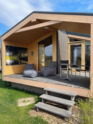 A modern wooden cabin with a covered porch, a large window with a view of a lake, and a dining area with outdoor furniture. - MoJo's Tiny House on the Elbe (Bleckede)