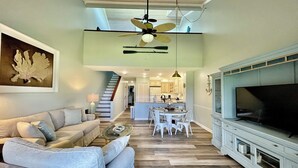 A spacious living room with a high ceiling fan, a large flat-screen TV, and a dining area with white chairs.