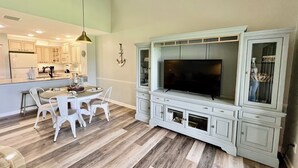 A modern kitchen with white cabinets, a dining table with chairs, a built-in TV, and a wooden floor.