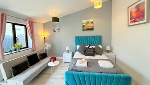 A modern bedroom with a teal upholstered bed, a grey sofa, a small white table, and a window with curtains. - 526A Farnham Rd  (Slough)