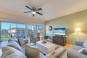 A spacious living room with a ceiling fan, a television, and a view of a building through large windows.