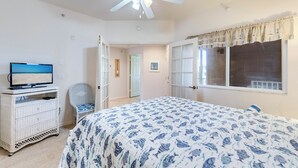 Un dormitorio con cama, televisión, ventilador de techo y ventana con cortinas.