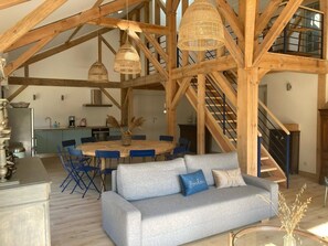 A spacious living area with a high ceiling, exposed wooden beams, a dining table with blue chairs, and a grey sofa with a blue pillow. - Barrail Estate – Large Landes House (20 beds) (Soustons)