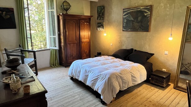Ein Schlafzimmer mit einem hölzernen Bett, einem Schreibtisch mit Schale und Tasse, einem grünen Vorhang und einem Fenster mit Blick auf Bäume.