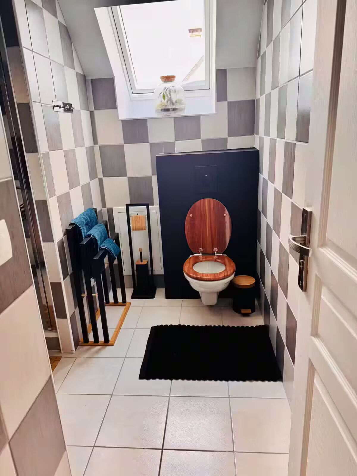A bathroom with a checkered tile wall, a skylight, a wooden toilet, and a black rug.