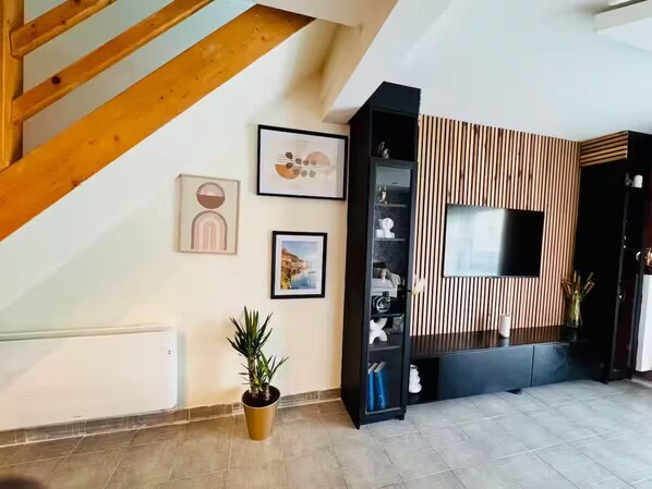 A modern living room with a wooden staircase, a flat-screen TV mounted on a wooden panel wall, and a potted plant.